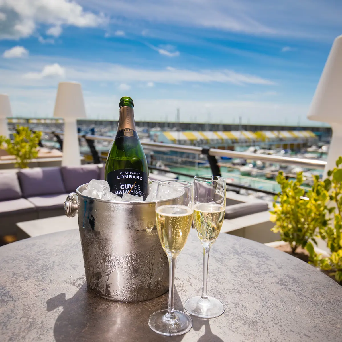 A bottle of champagne and two wine glasses on a table on a wooden deck terrace, with Brighton Marina in the background.